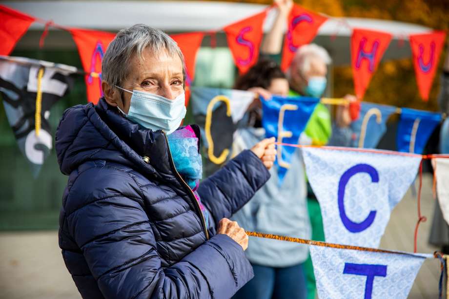 Eine Aktivistin der KlimaSeniorinnen hält zwei Protestbanner und schaut in die Kamera. Im Hintergrund sieht man weitere Demonstrant*innen. Sie stehen vorm Europäischen Gerichtshof für Menschenrechte in Straßburg, dessen Pforte im Hintergund verschwommen zu erkennen ist.