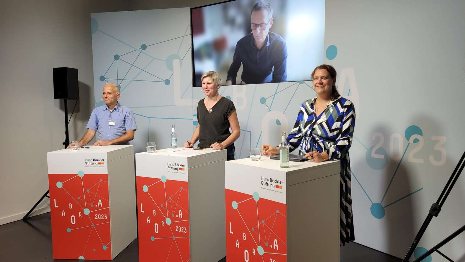 Dr. Immanuel Stieß (Links), Dr. Ulrike Spangenberg (Mitte) und Prof. Dr. Johanna Wenckebach (rechts) stehen hinter Redepulten auf einer Bühne. Über ihnen wird Dr. Peter Bleses digital auf einem Bildschirm eingeblendet.